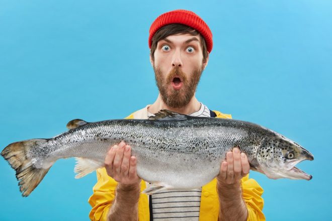 Indoor shot of astonished bearded fisherman dressed casually holding huge fish looking at camera with bugged eyes and jaw dropped being shocked to catch such big trout or salmon. Surprisment